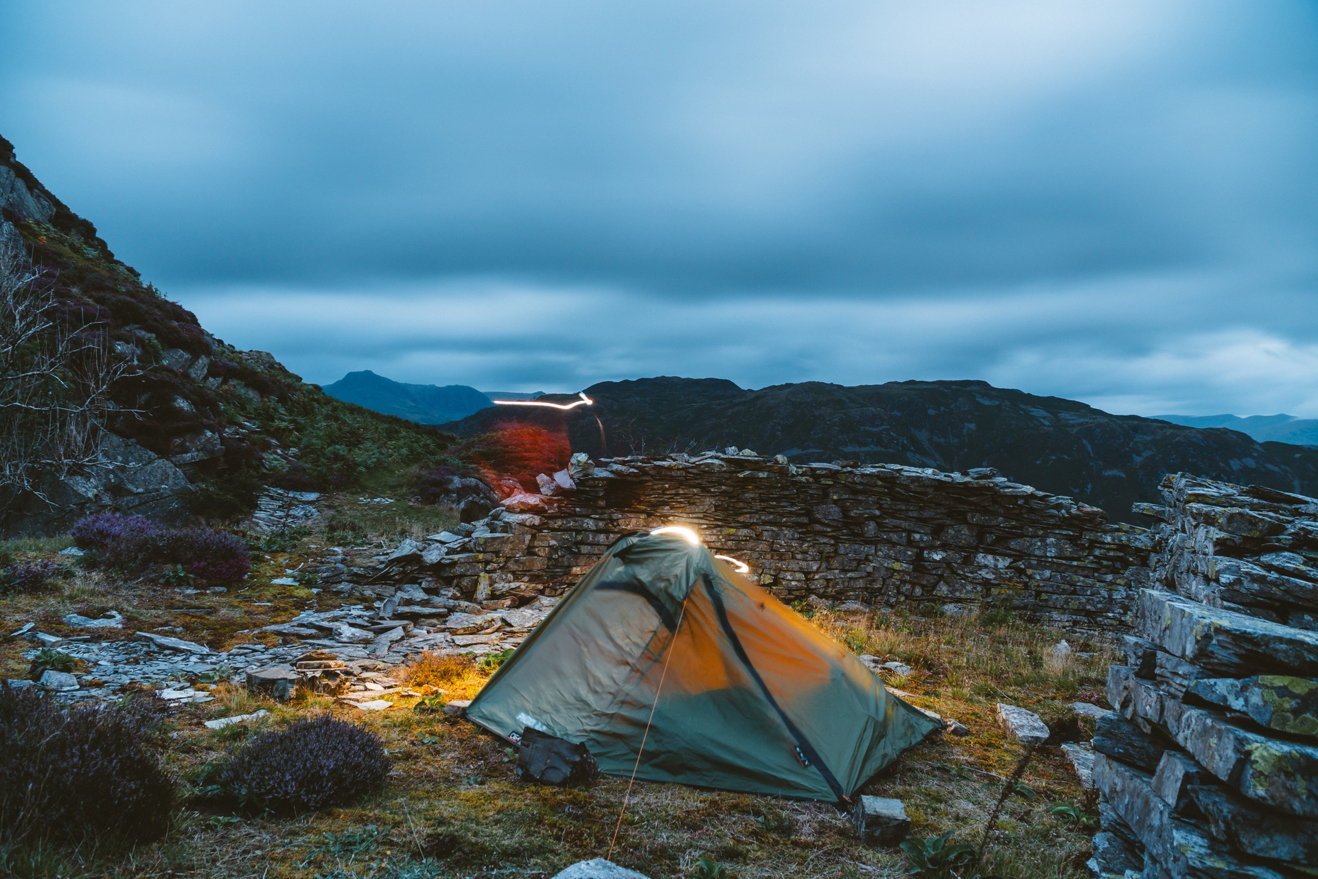 A glowing tent nestled in a rocky landscape under a twilight sky. Long exposure creates light trails.